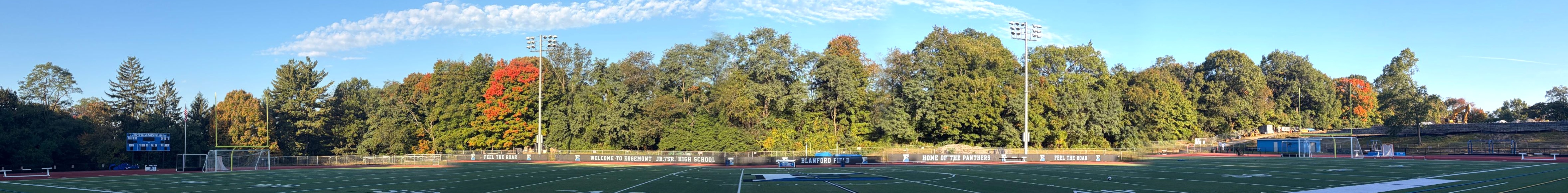 Panorama of the wooded ridge along the Edgemont High School track and Blanford Field, from the scoreboard end to the construction area, with fence banners reading Feel the Roar, Welcome to Edgemont Jr./Sr. High School, Blanford Field and Home of the Panthers. Two red lines mark the stretch of trees affected, from the trees just past the goalpost to the trees above the Welcome to Edgemont banner.